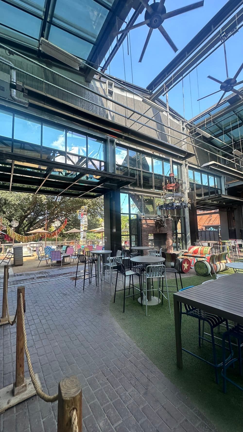 Outdoor dining area with modern tables, greenery, and large windows under a clear blue sky.