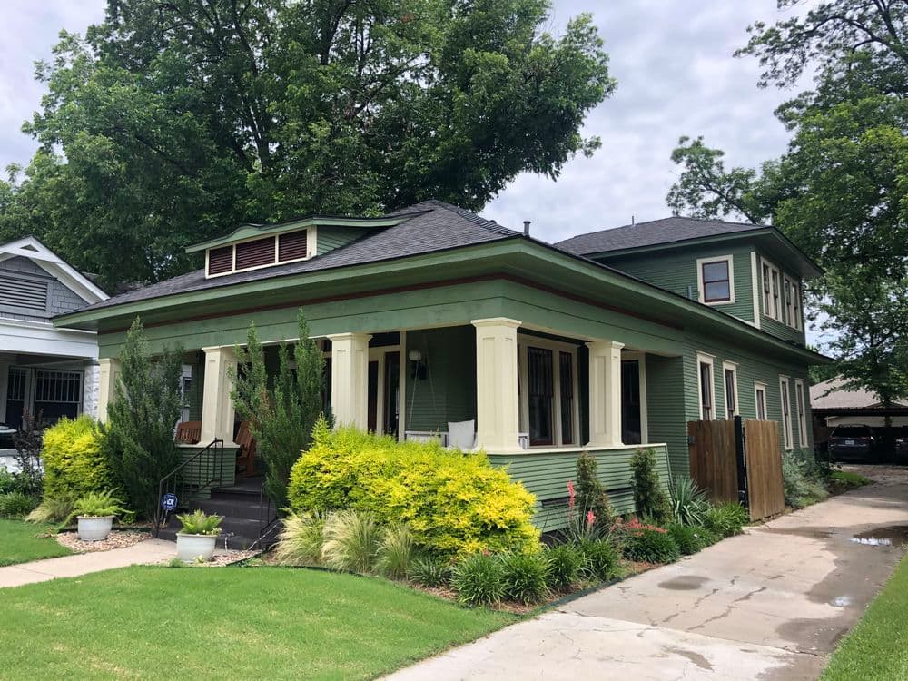 Green two-story house with porch, lush landscaping, and cloudy sky in the background.